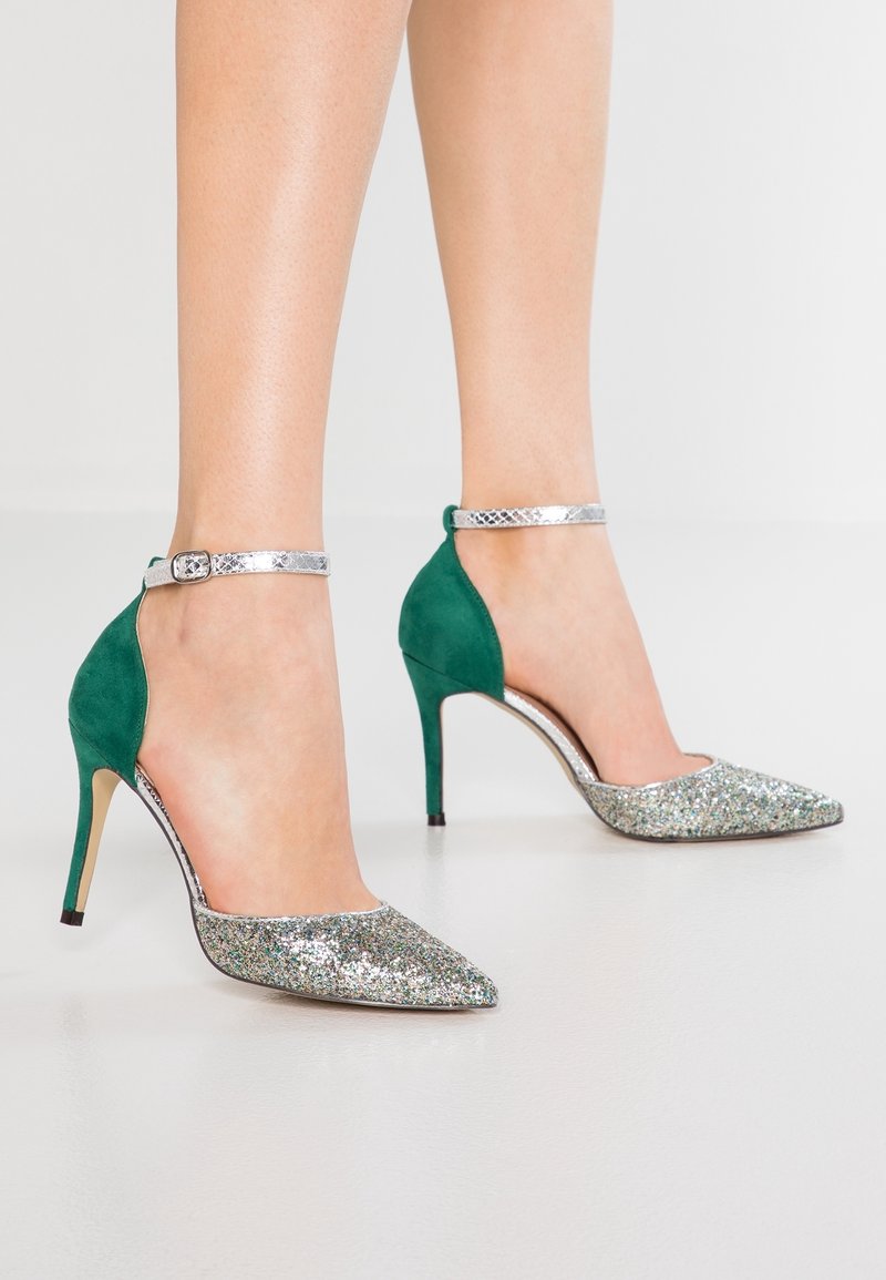 Femme portant des chaussures à talons hauts vertes et argentées avec des bouts pointus scintillants et des brides argentées à la cheville, debout sur un sol blanc.