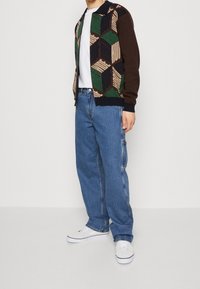 Brun och grönmönstrad bomberjacka kombinerad med blåa löst sittande jeans och vita sneakers. Tyget verkar mjukt med ribbade muddar.