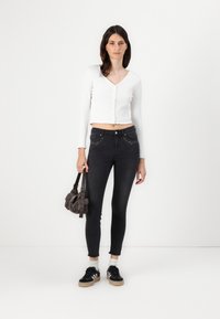 Cárdigan corto de canalé blanco con escote en V, combinado con jeans skinny negros de cintura alta con bolsillos adornados, y zapatillas negras.