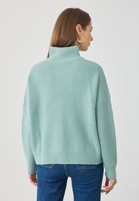 Miętowa zielona sweter z prążkowanym wzorem, o wysokim kołnierzu i opadających ramionach, zestawiony z ciemnoniebieskimi dżinsami. Teksturowany splot i luźny krój.