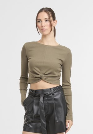Donna con i capelli raccolti, che indossa un crop top verde oliva a spirale e dei pantaloni corti di pelle nera a vita alta con una cintura, in piedi su uno sfondo bianco.