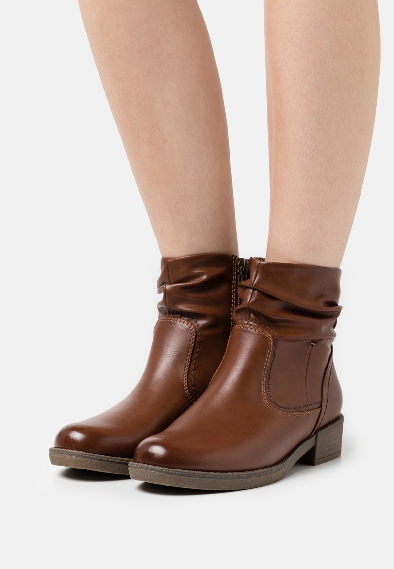 Bottines en cuir marron avec petits talons épais portées sur jambes nues, présentées sur un fond blanc.