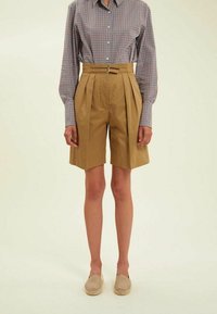 Shorts plissés beiges à taille haute avec passants de ceinture, associés à une chemise à manches longues à carreaux bleus et rouges. Texture de tissu lisse.