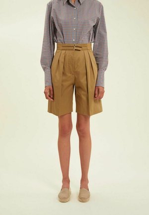 Shorts plissés beiges à taille haute avec passants de ceinture, associés à une chemise à manches longues à carreaux bleus et rouges. Texture de tissu lisse.