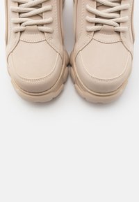 Beiga sportskor har en slät mockaliknande ovandel, snörning framtill och tjocka vadderade sålar. Diskreta sömmar framhäver designen.