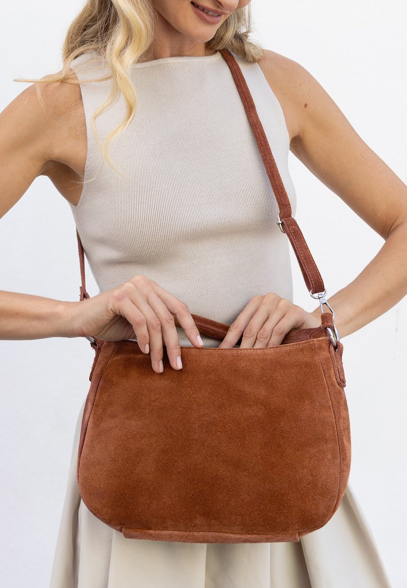 Femme en haut beige sans manches et jupe, portant la main dans un sac à bandoulière en daim marron de taille moyenne avec une sangle réglable.