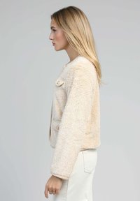 Jeune femme aux longs cheveux blonds portant une veste crème texturée et un pantalon blanc, se tenant de profil contre un fond uni.