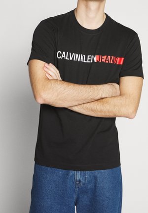 Person mit verschränkten Armen, trägt ein schwarzes T-Shirt mit dem weißen und roten Schriftzug „CALVINKLEIN JEANS“ und blaue Jeans.