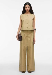 Kvinde med langt mørkt hår iført en ærmeløs beige knap-op top og brede beige bukser, der holder en mørk clutch taske og bærer brune sko.