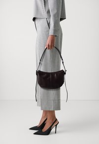 Borsa in pelle marrone scuro con forma curvata e manico corto, tenuta in una mano. L'abbigliamento presenta un motivo a scacchi in bianco e nero.
