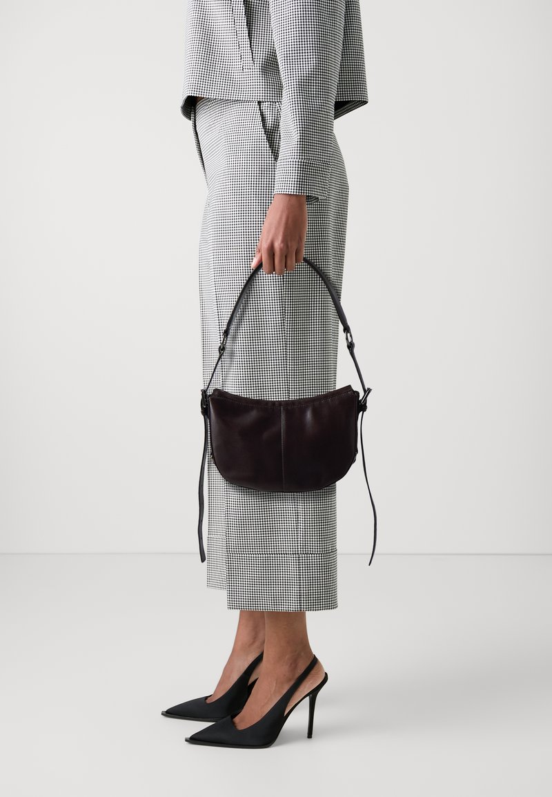 Borsa in pelle marrone scuro con forma curvata e manico corto, tenuta in una mano. L'abbigliamento presenta un motivo a scacchi in bianco e nero.