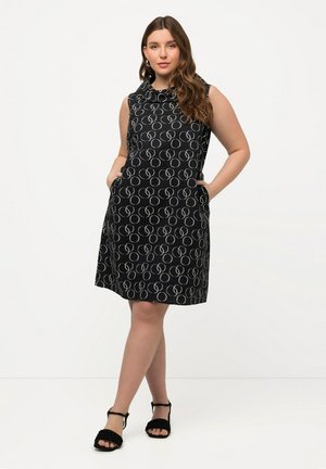 Schwarzes ärmelloses Kleid mit einem kreisförmigen Muster in Hellgrau, ausgestattet mit zwei Seitentaschen und einem lässigen Schnitt. Kombiniert mit schwarzen Sandalen mit Riemchen.