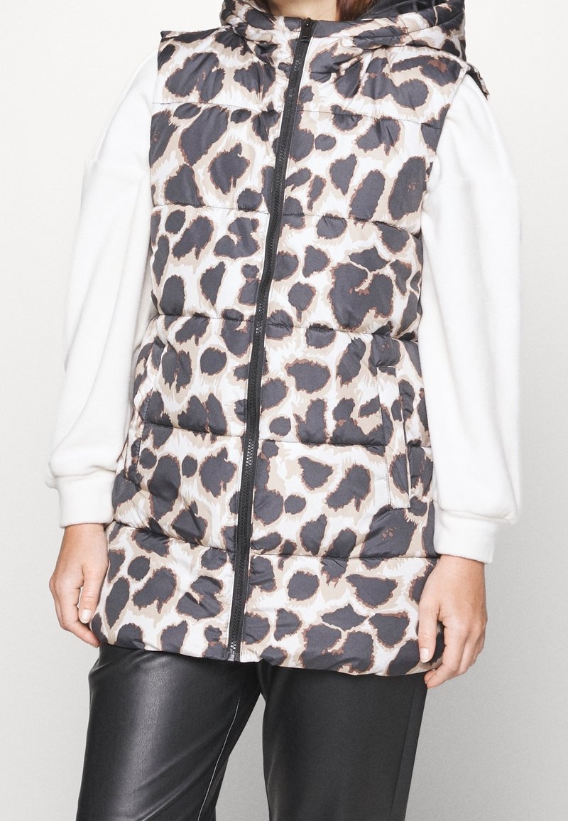 Mujer con chaleco acolchado sin mangas con capucha y estampado de leopardo en negro y beige, sobre una camisa blanca de manga larga y pantalones negros.