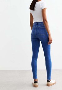 Mujer con jeans skinny azul de cintura alta, camiseta blanca ajustada de manga corta y zapatillas beige, de pie de espaldas sobre un fondo neutro.
