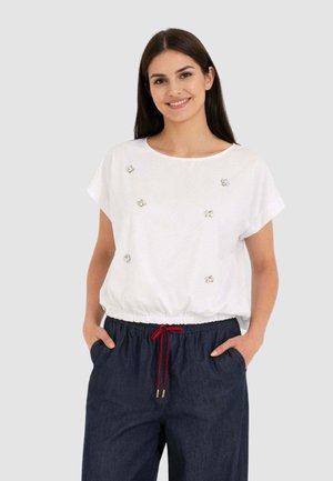 Donna sorridente che indossa una maglietta bianca a maniche corte con piccole decorazioni e pantaloni blu scuro con coulisse rossa, mani nelle tasche.