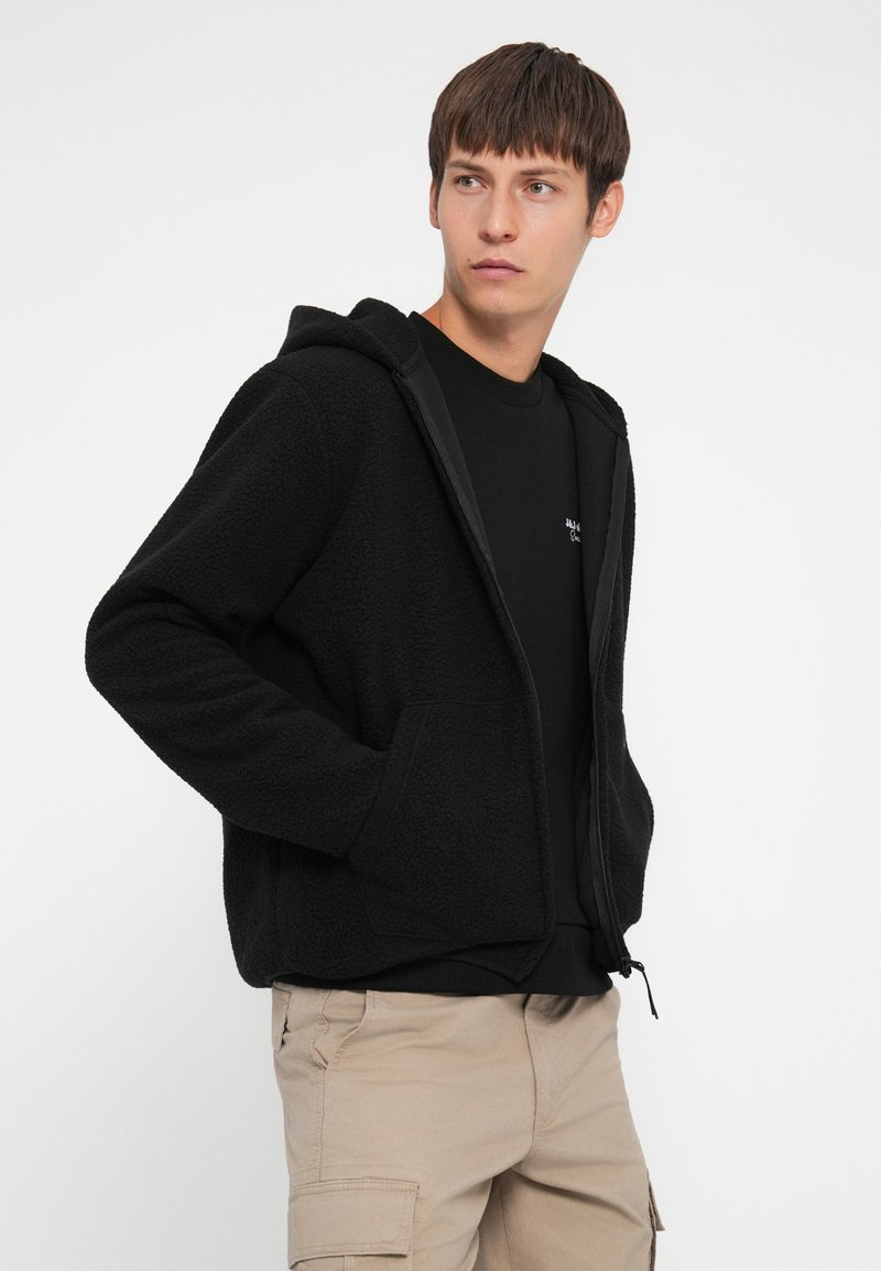 Felpa nera in pile con tasche frontali, chiusura a zip completa, tessuto testurizzato e cappuccio integrato, indossata sopra una maglietta nera e pantaloni beige.