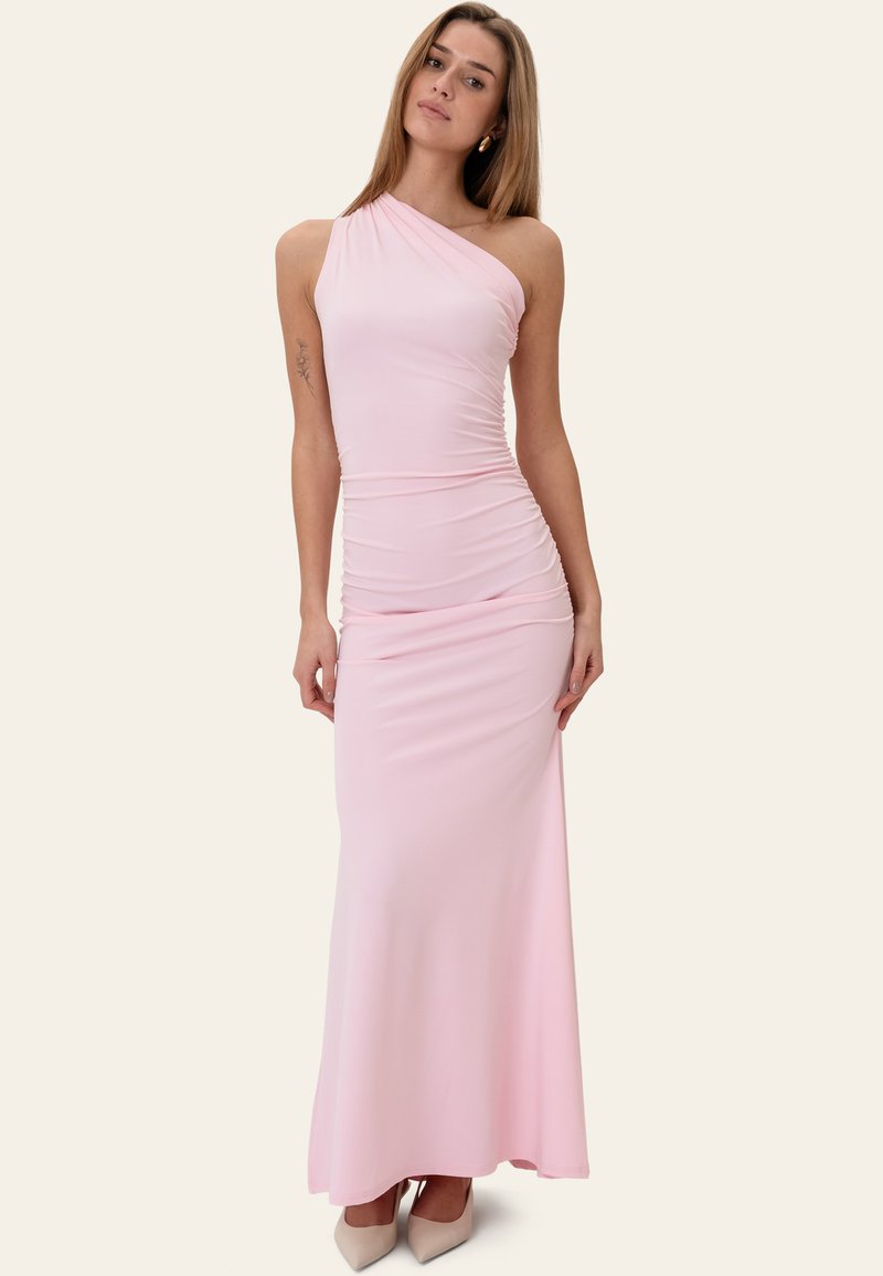 Femme portant une robe longue rose pâle à une épaule froncée avec des talons beiges pointus devant un fond uni.