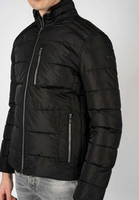 Giacca a vento nera con colletto alto, caratterizzata da una trama a costine orizzontali, con tasche laterali e sul petto con zip, e materiale esterno liscio.
