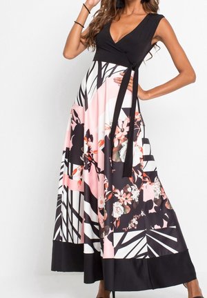 Femme portant une robe noire sans manches à effet portefeuille avec une jupe fluide à motifs géométriques et floraux en rose, blanc et noir, posant avec la main sur la hanche.