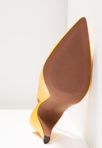 Chaussure à talon haut jaune avec bout pointu et accent noir à l'extrémité. Matériau en cuir lisse avec une semelle marron texturée.