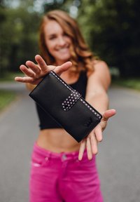 Black wallet with a pink polka dot strap, featuring a logo embossed on the front. Smooth texture, rectangular shape, and snap closure.