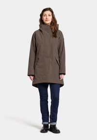 Bruine, lange parka met een hoge kraag, zijzakken en een rechte snit. Gekocht over marineblauwe jeans en zwarte enkellaarsjes. Soepele stofstructuur.