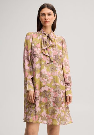 Frau trägt ein langärmeliges Kleid mit pinken Blumen auf grünem Hintergrund und eine gebundene Schleife am Ausschnitt.