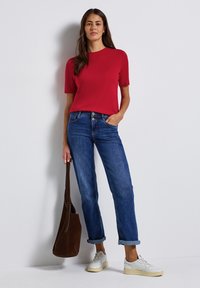 Röd kortärmad skjorta matchad med blå denimjeans, uppvikta ärmslut, och vita sneakers. Modellen håller en brun mockaväskan.