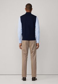 Mornarski pleteni prsluk preko svijetloplavog dugog rukava, u kombinaciji s bež chinos hlačama i tamnim kožnim cipelama. Pogled s leđa otkriva strukturu odjevnog predmeta.