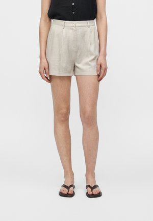 Femme portant un short beige taille haute sur mesure, un top noir sans manches boutonné et des sandales noires type tongs, debout devant un fond clair uni.