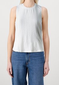 Donna che indossa un top senza maniche in pizzo bianco e jeans a vita alta blu, in piedi con le braccia rilassate lungo i fianchi su sfondo bianco.