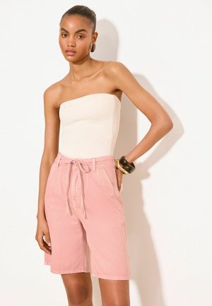Femme portant un haut blanc sans bretelles, un short rose taille haute avec cordon, de grandes boucles d'oreilles et des bracelets épais à un poignet.