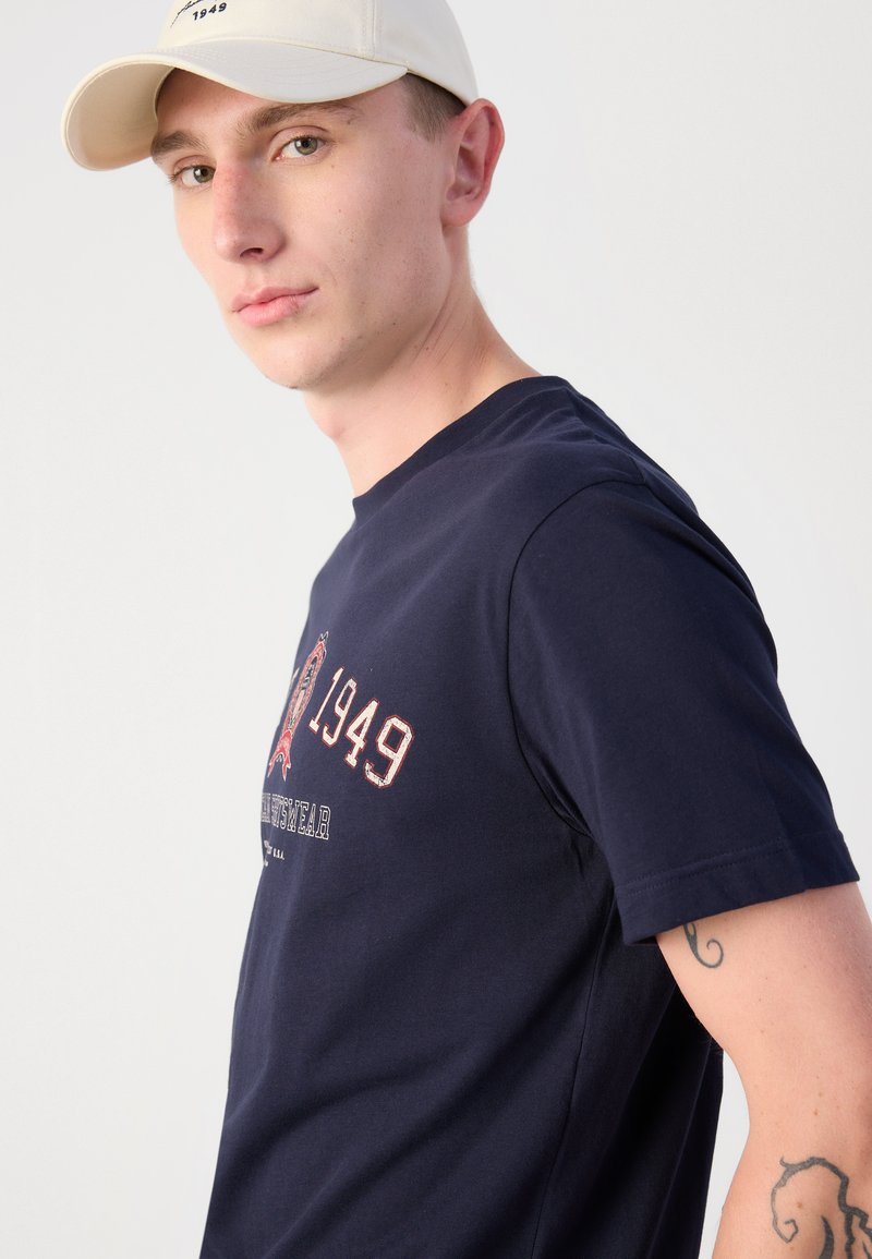 Granatowa bawełniana koszulka z krótkim rękawem, z grafiką na przedzie, na której widnieje tekst "1949" oraz elementy dekoracyjne, gładka struktura.