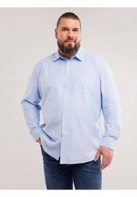 Bärtiger Mann mit zurückgebundenen Haaren, der ein hellblaues Hemd mit Knöpfen und eine Jeans trägt, steht mit einer Hand in der Tasche.