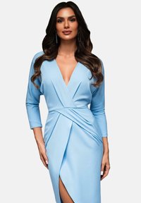 Abito azzurro chiaro con maniche lunghe, profondo scollo a V, design plissé e spacco fino alla coscia. Tessuto liscio e silhouette aderente.