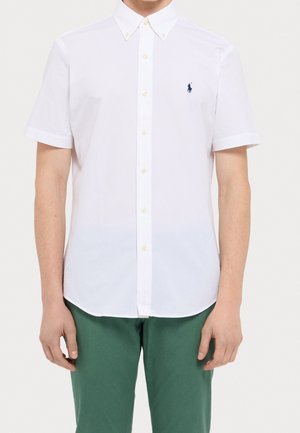 Mężczyzna ubrany w białą koszulę z krótkim rękawem zapinaną na guziki z małym ciemnym logo na piersi i zielone spodnie, stojący na jednolitym tle.