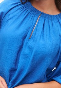 Blusa azul con un escote fruncido, una apertura en forma de lágrima con cierre de botón, y tela texturizada. Las mangas cortas completan el diseño.