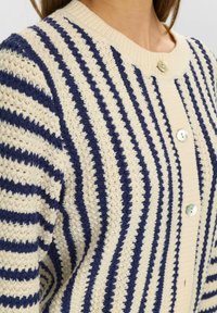 Gestreifte Strickjacke in Creme und Marineblau, mit einem strukturierten Strickdesign, ribbeligem Kragen und Bündchen sowie dekorativen Knöpfen auf der Vorderseite.