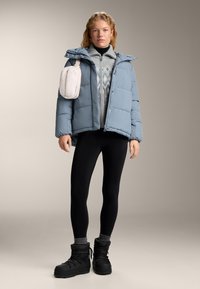 Modrá prošívaná bunda s kapucí, šedý vzorovaný svetr, černé legíny a černé zimní boty. Součástí je malá krémová taška připevněná.