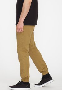 Beżowe, tapered spodnie z elastycznymi mankietami, bocznymi kieszeniami i gładką teksturą, zestawione z czarnym obuwiem i czarnym T-shirtem.