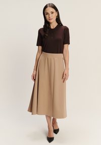 Hnedé polo tričko s krátkymi rukávmi a tromi gombíkmi, skombinované s béžovou midi sukňou vo forme A. Modelka má na sebe čierne lodičky s špicatou špičkou.