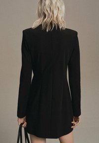 Vestido blazer negro con mangas largas, corte entallado, textura suave y una sola costura trasera. Acompañado de un bolso negro delgado.