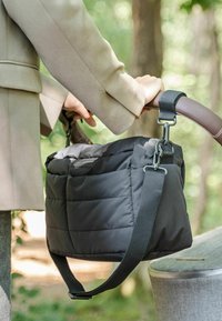 Sort quiltet tote taske med et gråt stofinteriør, justerbar skulderrem og brunt læderhåndtag, præsenteret i en naturlig setting.