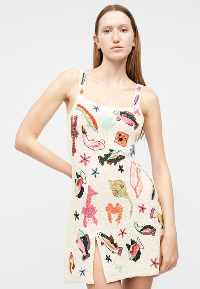 Νέα γυναίκα με ίσια μαλλιά, που φοράει λευκό αμάνικο φόρεμα διακοσμημένο με πολύχρωμα κεντημένα θαλάσσια πλάσματα και με σκίσιμο μπροστά.