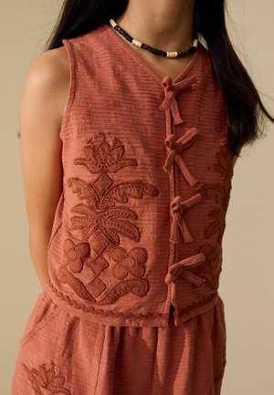 Top fără mâneci, de culoare ruginie, cu broderie florală și legături frontale nodate, purtat cu partea de jos asortată și un colier din mărgele alb-negru.