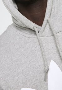 Grå sweatshirt med strukturerad yta, med dragsko i huvan och en stor vit logotyp på framsidan.