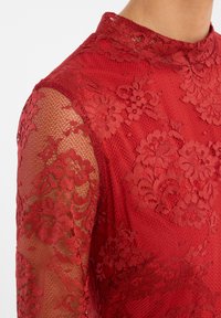 Haut en dentelle rouge avec un col montant et des manches transparentes à motifs floraux, présentant une texture et des superpositions délicates.