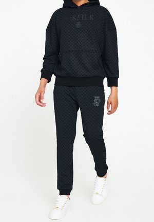 Persoon die een zwart patroon hoodie en bijpassende joggingbroek draagt met witte sneakers, poserend tegen een effen witte achtergrond.