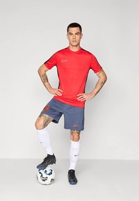 Punane Nike spordisärk mustade aktsentidega; sinised lühikesed püksid; valged sokid; mustad jalgpalliketsid; seisab jalgpallil; nähtavad tätoveeringud.