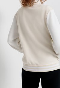 Jasno beżowa bomberka z białymi rękawami, ściągaczami przy mankietach i paskowanym dołem. Gładka tekstura i swobodny fason z luźnym krojem.